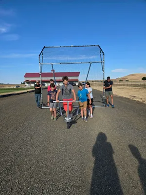 Youth and leaders wheeling the new backstop down a gravel path toward the field